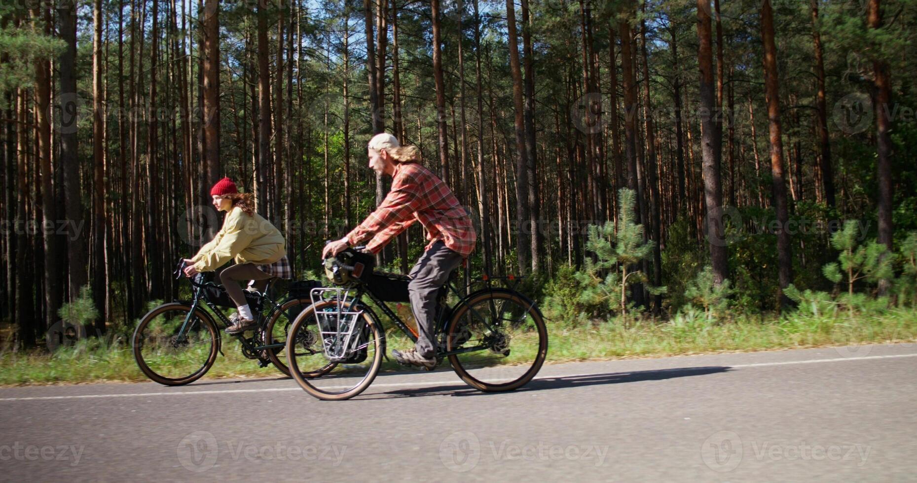 A young man and a woman ride bicycles along the road along the forest, side view photo