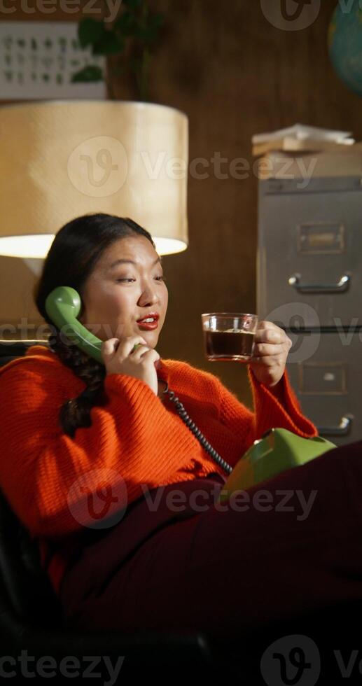 A woman is talking on a wired phone and drinking coffee sitting in a retro room. The camera zooms on her face photo