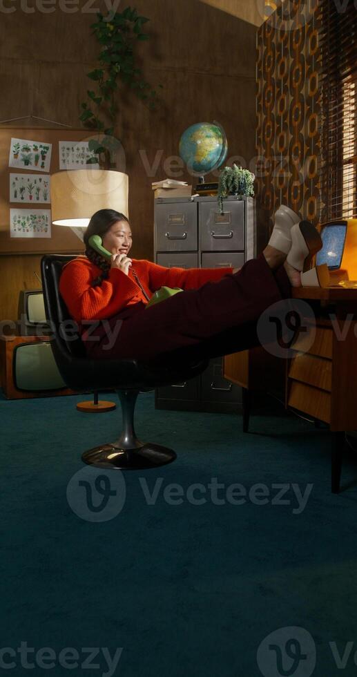 A woman is talking on a wired phone and drinking coffee sitting in a retro room. Her feet are on the desktop photo