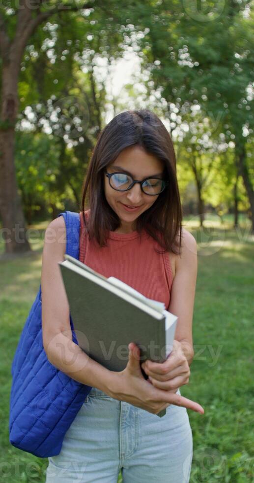 A young woman with glasses is smiling at the camera while standing in a city park. She holds a notebook and a book in her hands. Summer day photo