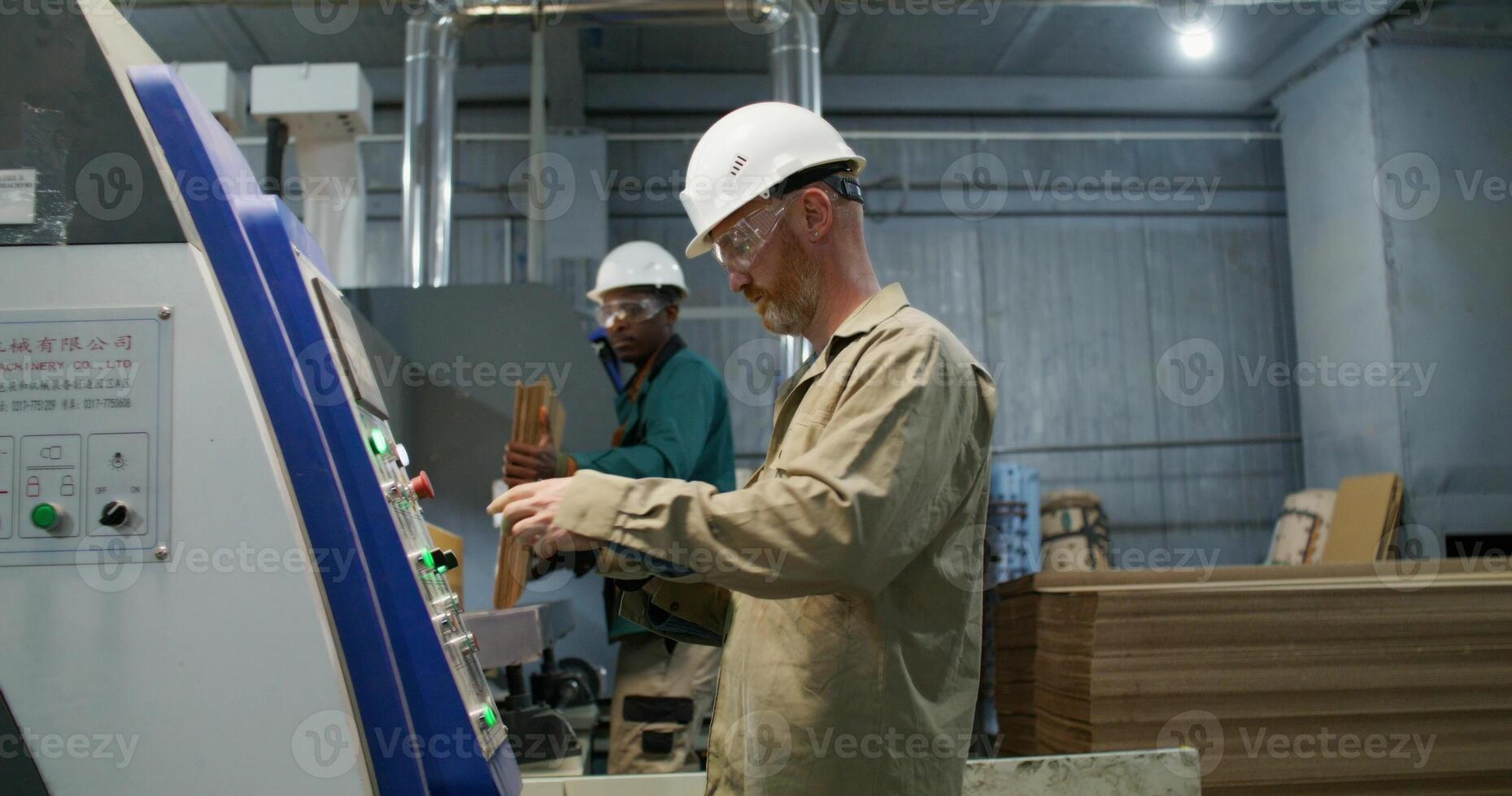 Two men work in a paper mill. One of them uses a tablet to control the operation of the machine. The second man brings stacks of cardboard to the machine photo