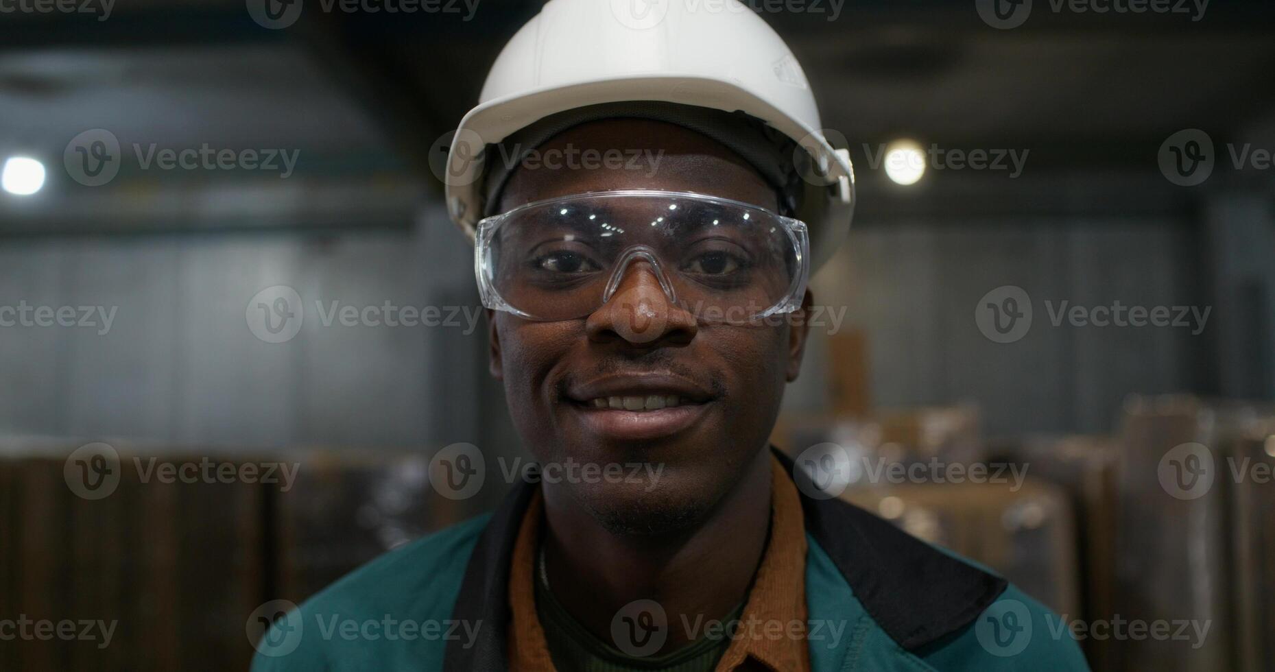A worker in a hard hat and safety glasses smiles at the camera standing at the factory. Close-up of his face. photo