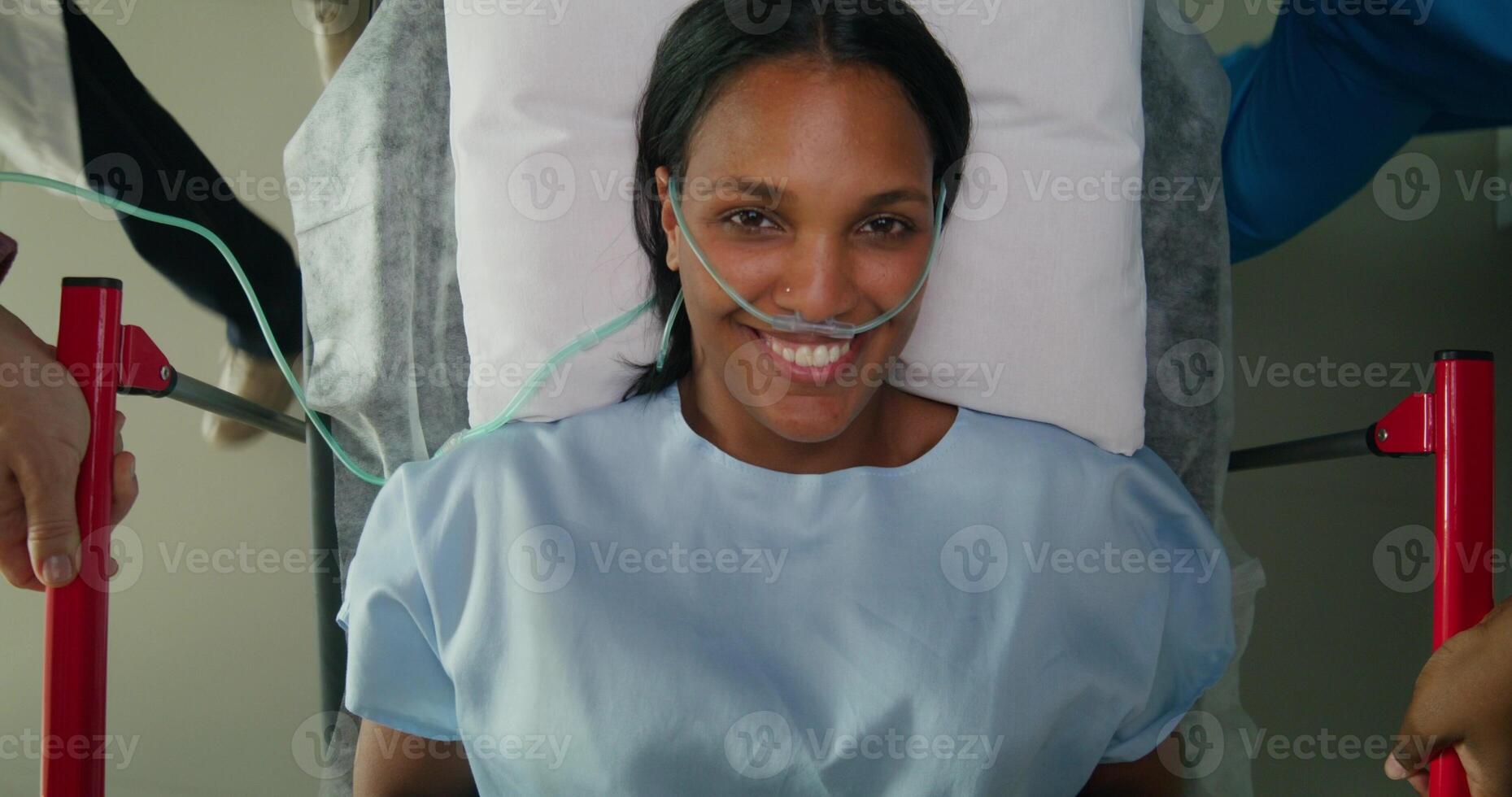 A woman with an oxygen tube in her nose smiles at the camera while lying on a medical gurney. She is being driven down the hospital corridor photo