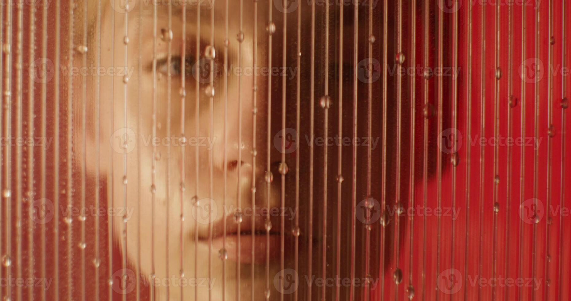 A woman looks at the camera with a serious look standing behind a glass with water running down about it in the studio. Close-up of her face photo