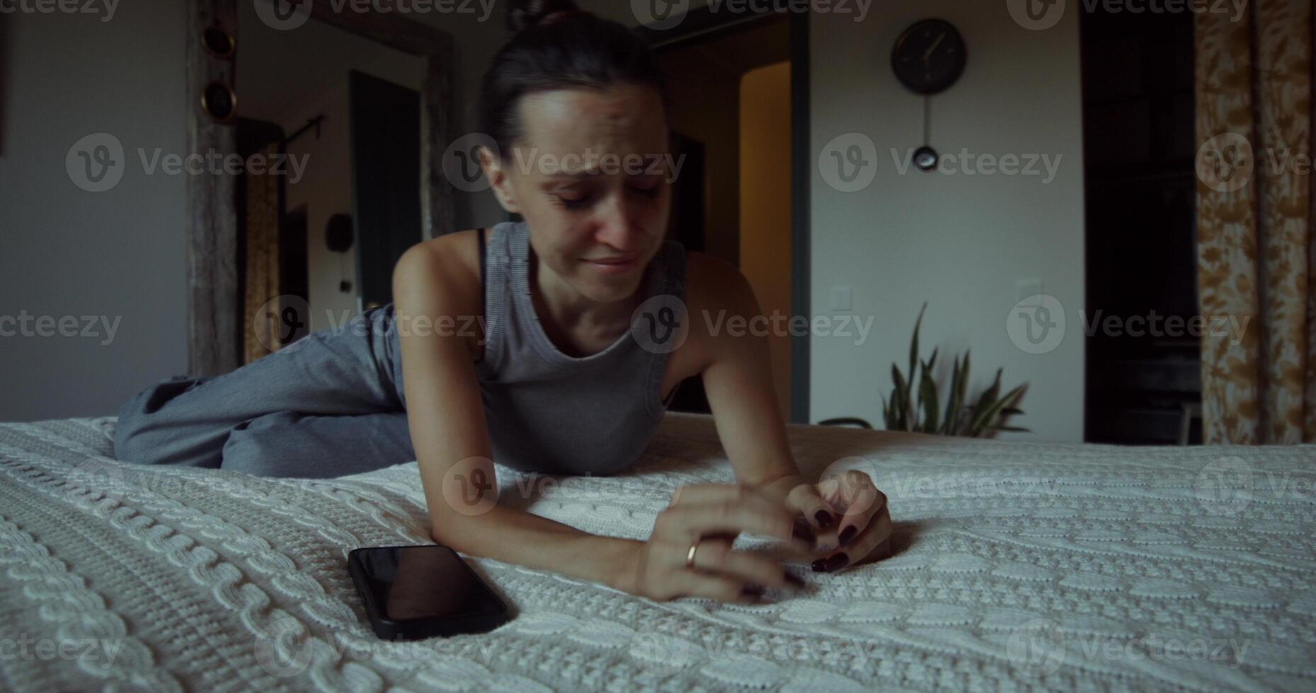 A woman is crying lying on a bed at home. She takes off her wedding ring and throws it away. Her mascara was leaking. The mobile phone is on the bed next to her photo