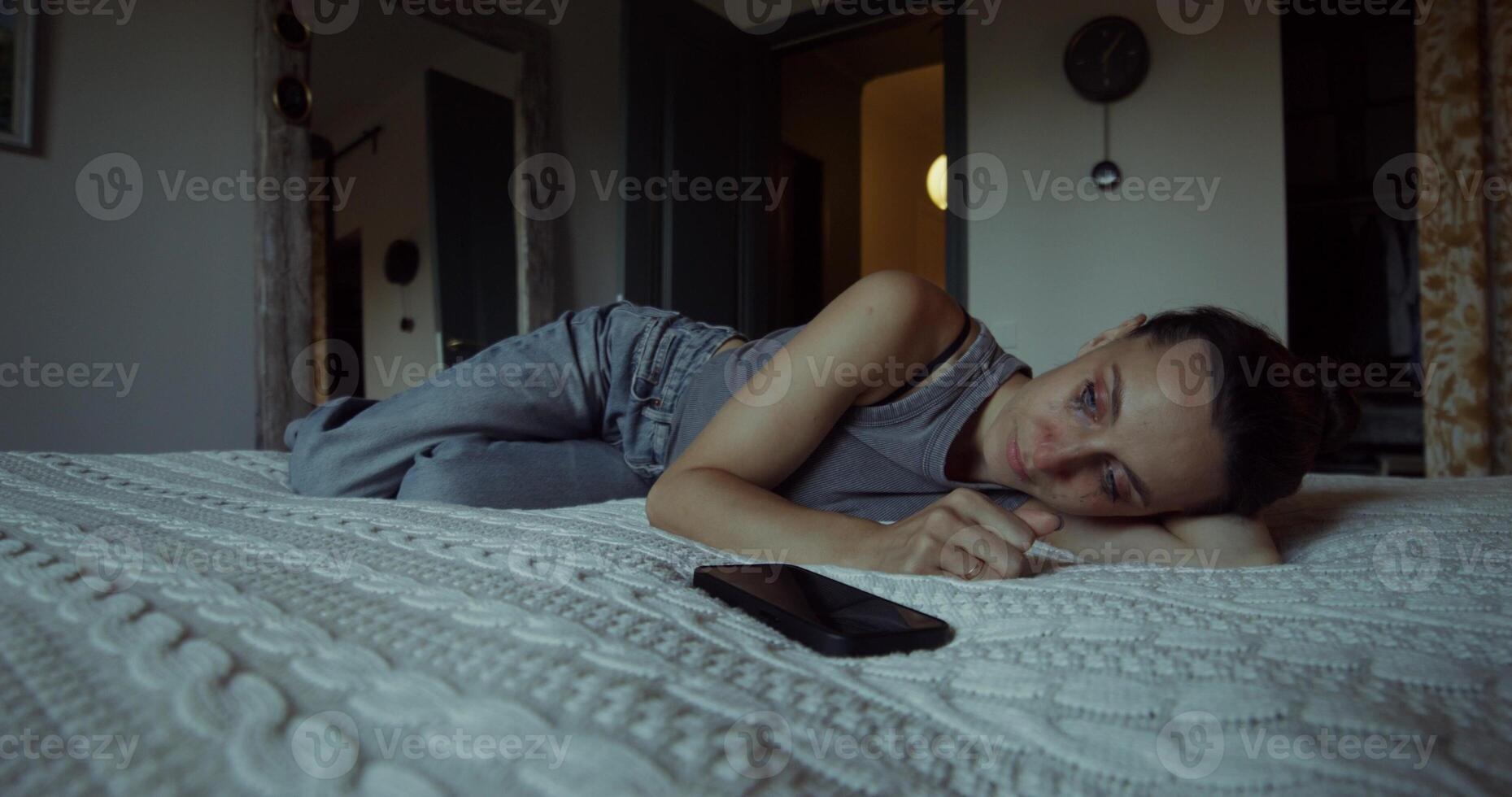 A woman is crying lying on a bed at home. Her mascara was leaking. The mobile phone is on the bed next to her photo