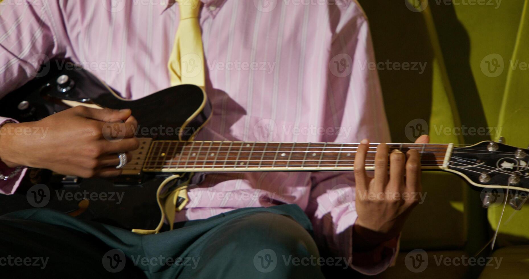 The man is smiling while playing the guitar. He is sitting on a sofa in a bright eighties interior photo