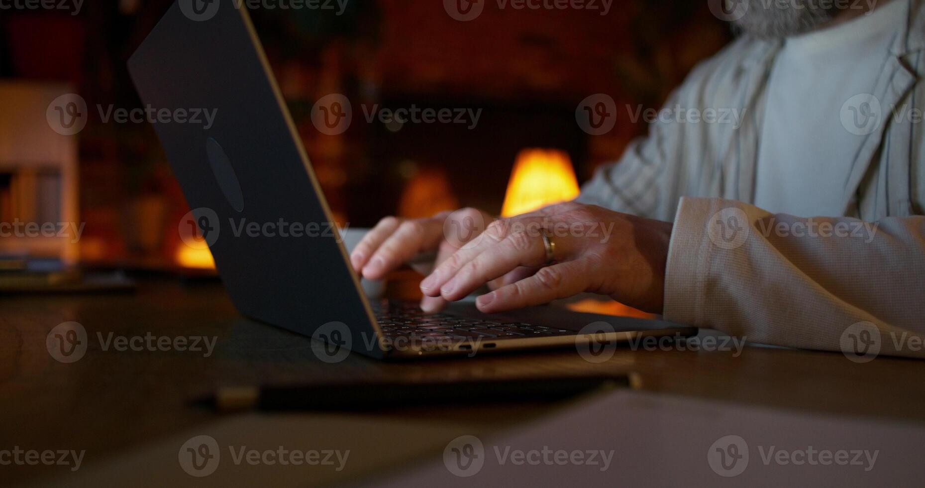 A man typing on a laptop sitting at a desk at night at home. Close-up of his hands, unrecognizable persona photo
