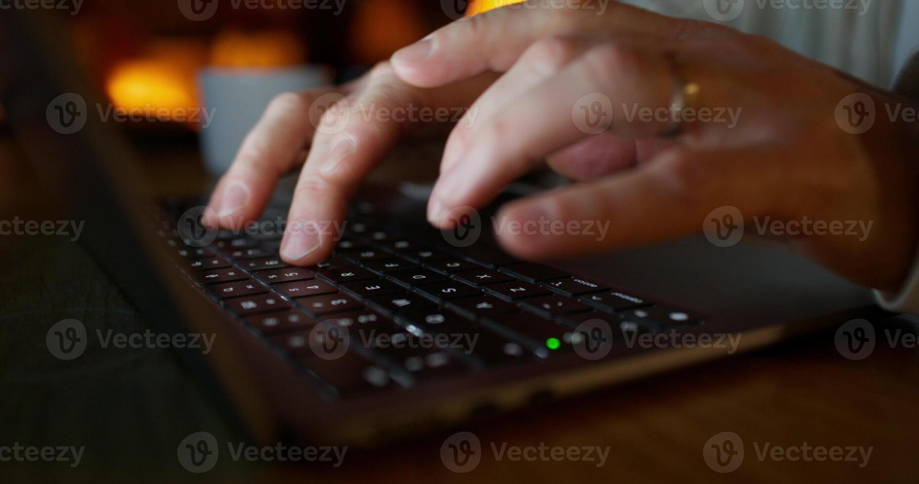 A man typing on a laptop while sitting at a desk at night. Close-up of his hands, unrecognizable persona photo