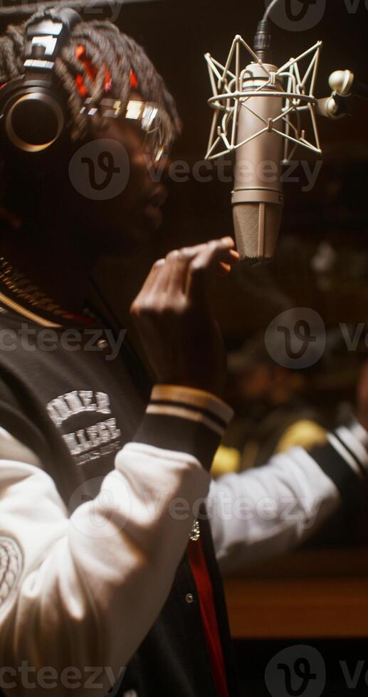 The rapper is recording a song in a recording studio. The sound engineer is sitting in the control room behind the glass photo