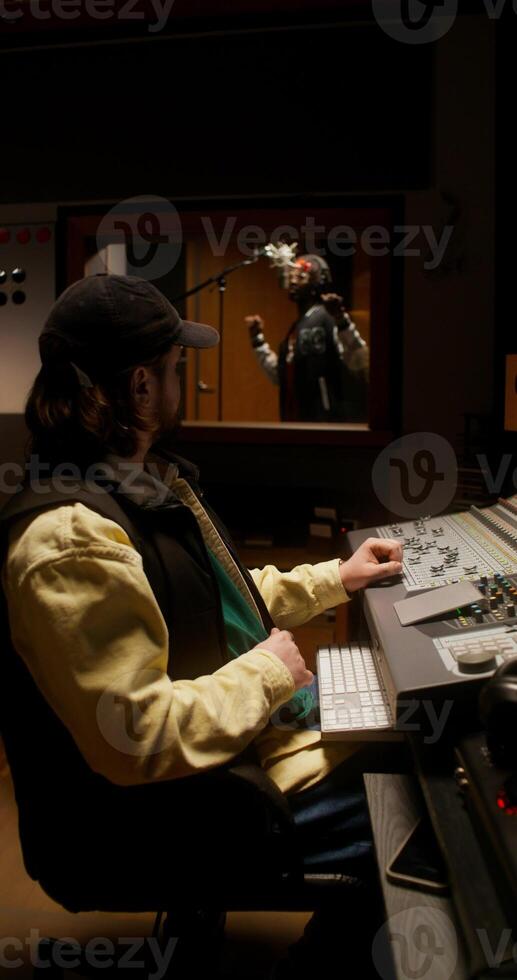 The sound engineer controls the recording process of the song in the studio. The rapper sings in the room behind the glass photo