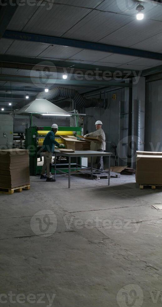 Two men in hard hats and uniforms use a cardboard cutting machine. Working in a paper mill photo