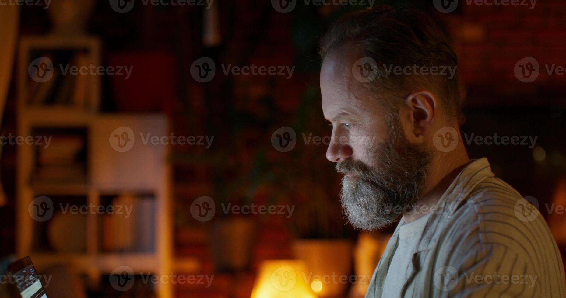 A man uses a laptop and drinks coffee sitting at a table at home. He looks focused or tired. Night work photo