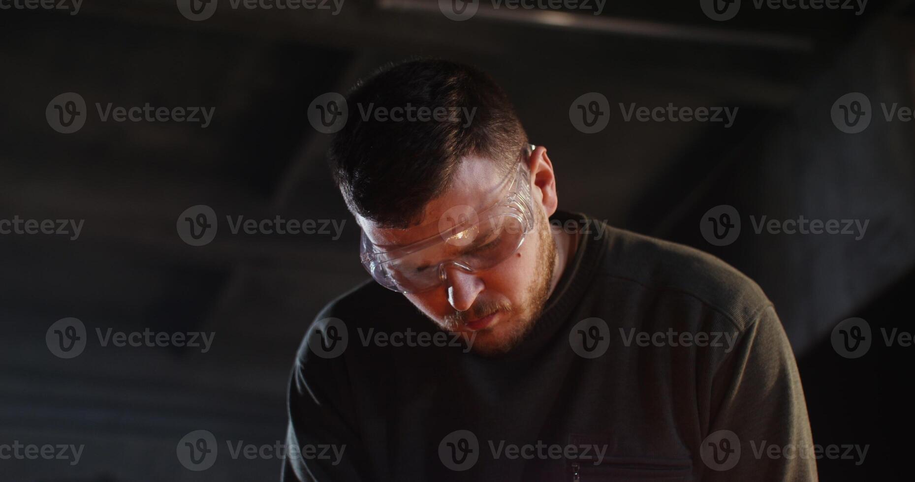 A man dressed in work clothes and goggles with a focused look uses a plasma cutter while working in a forge. A close-up of a man's face among flying metallic sparks. 4k, red komodo photo