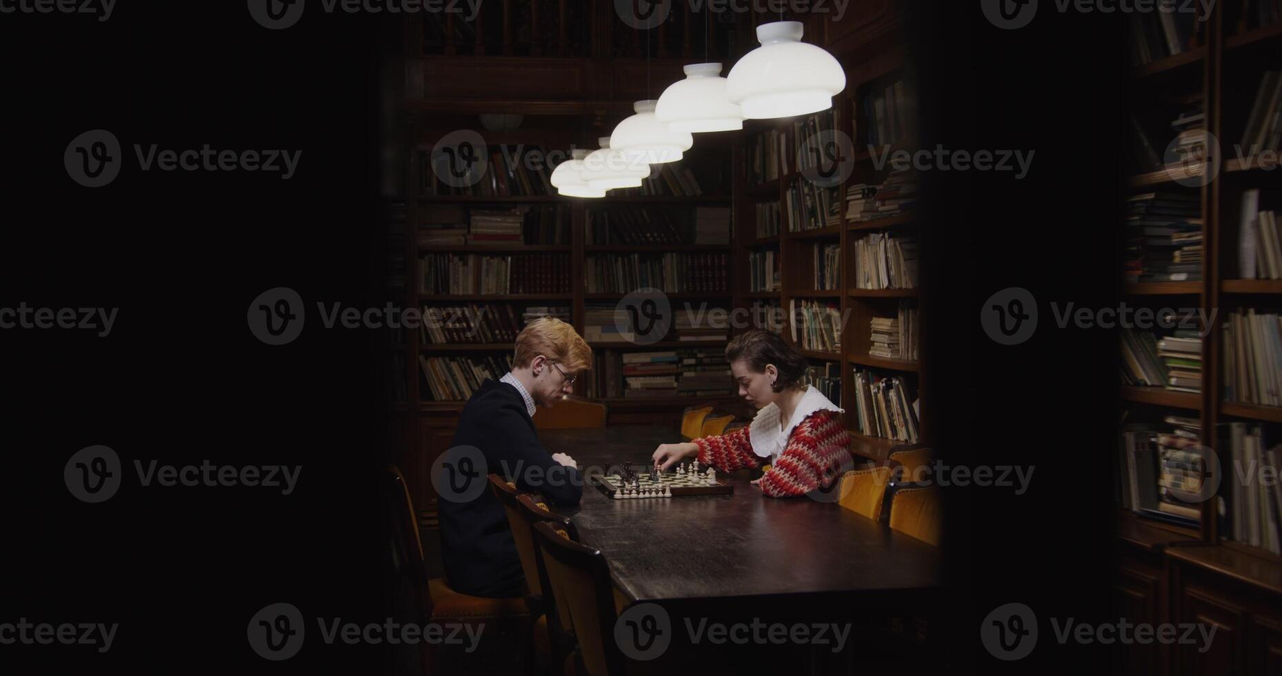 Young man and woman of European appearance start a chess game, thinking intently, sitting opposite each other at a table in the library photo