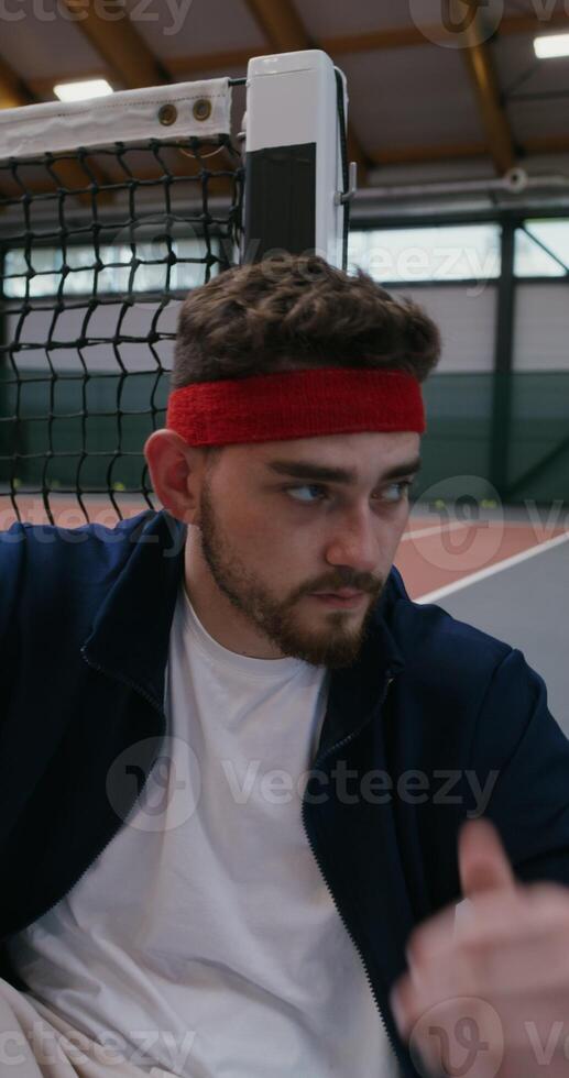 A young man of European appearance is resting after training, leaning his back and head on a tennis net stretched in the middle of the court, close-up, vertical photo