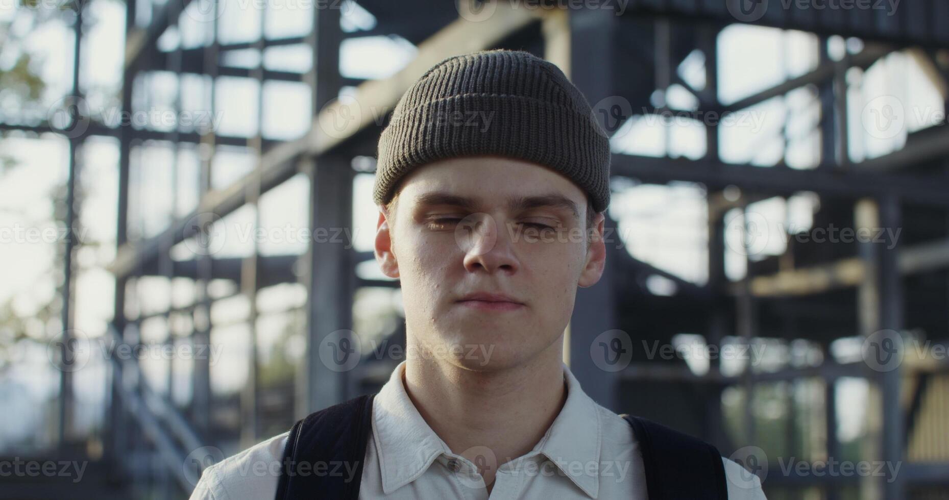 A young man of European appearance in a knitted hat with a backpack on his back looks straight into the camera with a serious look, portrait photo