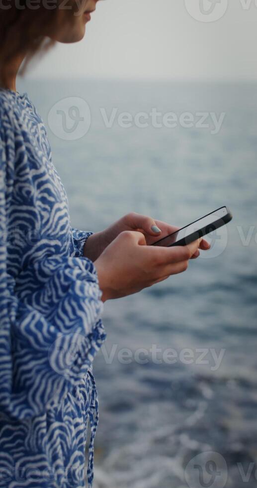 A woman is typing on a mobile phone while standing on the rocks on the seashore. No face, vertical . The woman is blurry. 4k , red komodo photo