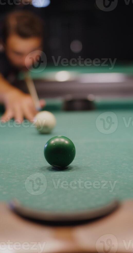 A young man of European appearance intently aims and hits a billiard ball with a cue, driving it into the hole, vertical . photo