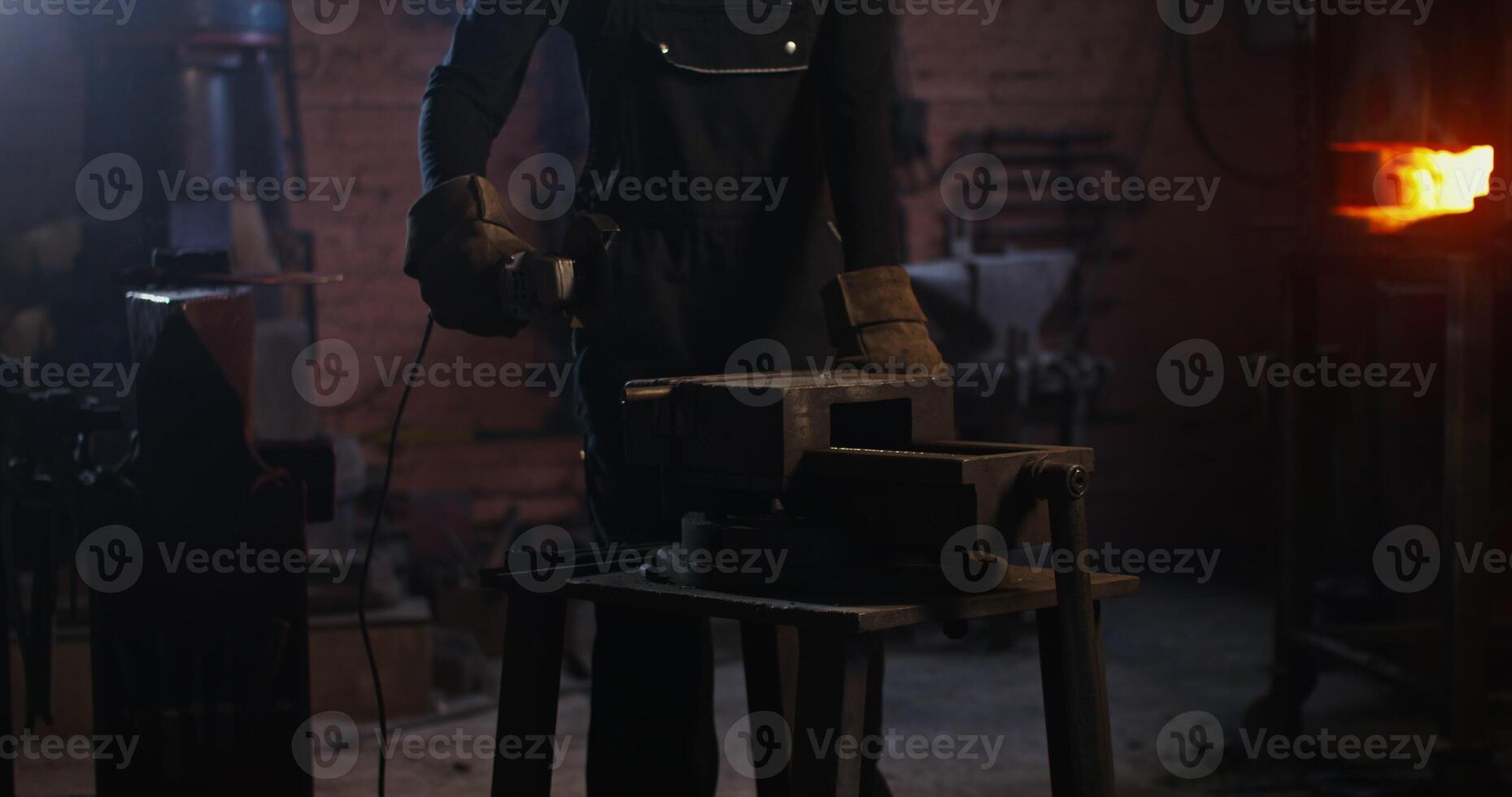 A blacksmith of African American appearance in a leather apron and gloves cuts off a piece of metal workpiece using an angle grinder. close-up, no face. 4k, red komodo photo