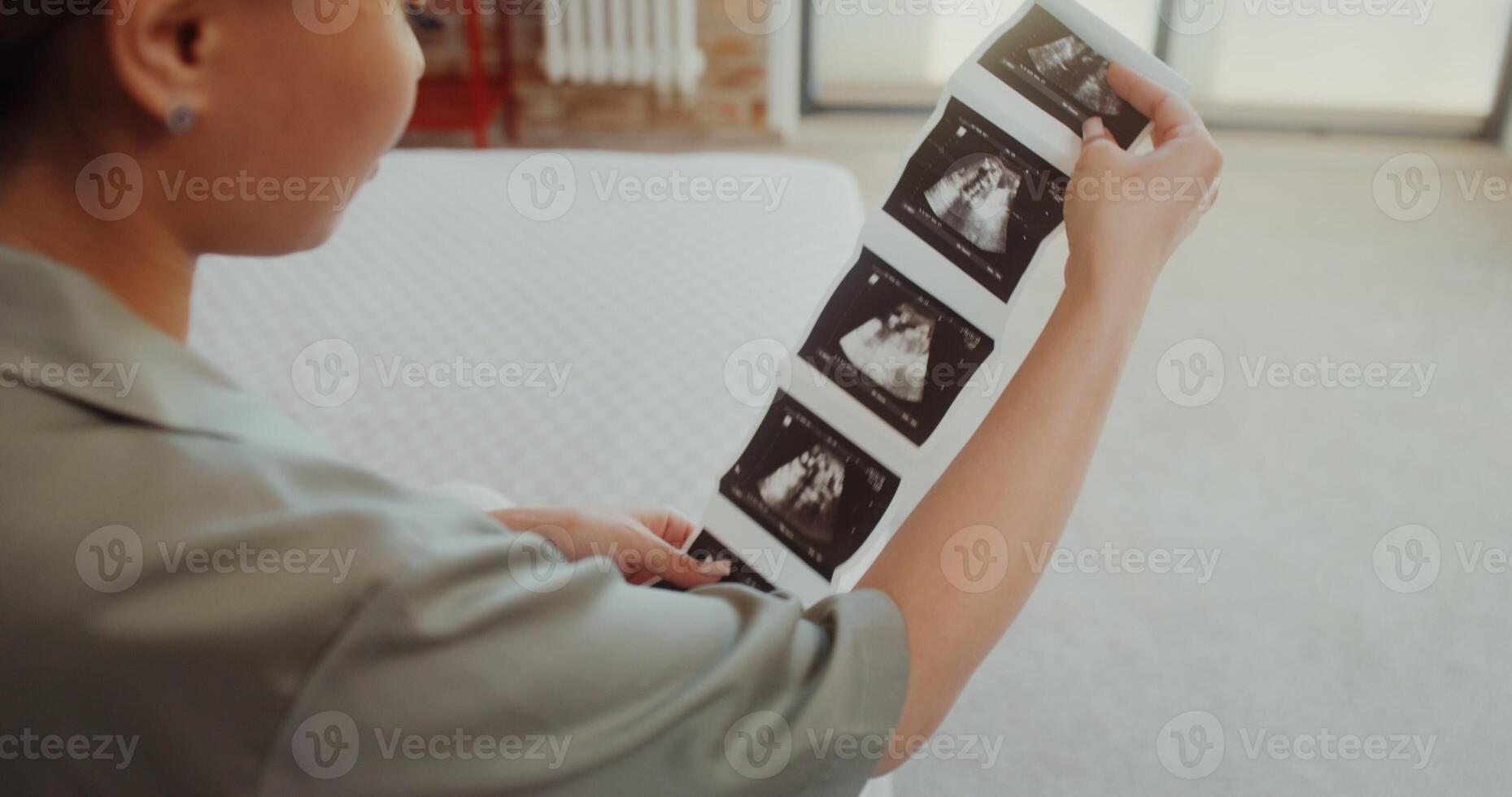 A woman examines ultrasound images sitting on a bed at home, view from over her shoulder photo