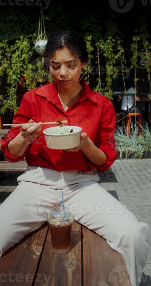 A young woman in casual clothes eats Asian food using chopsticks while sitting on a bench on a city street photo