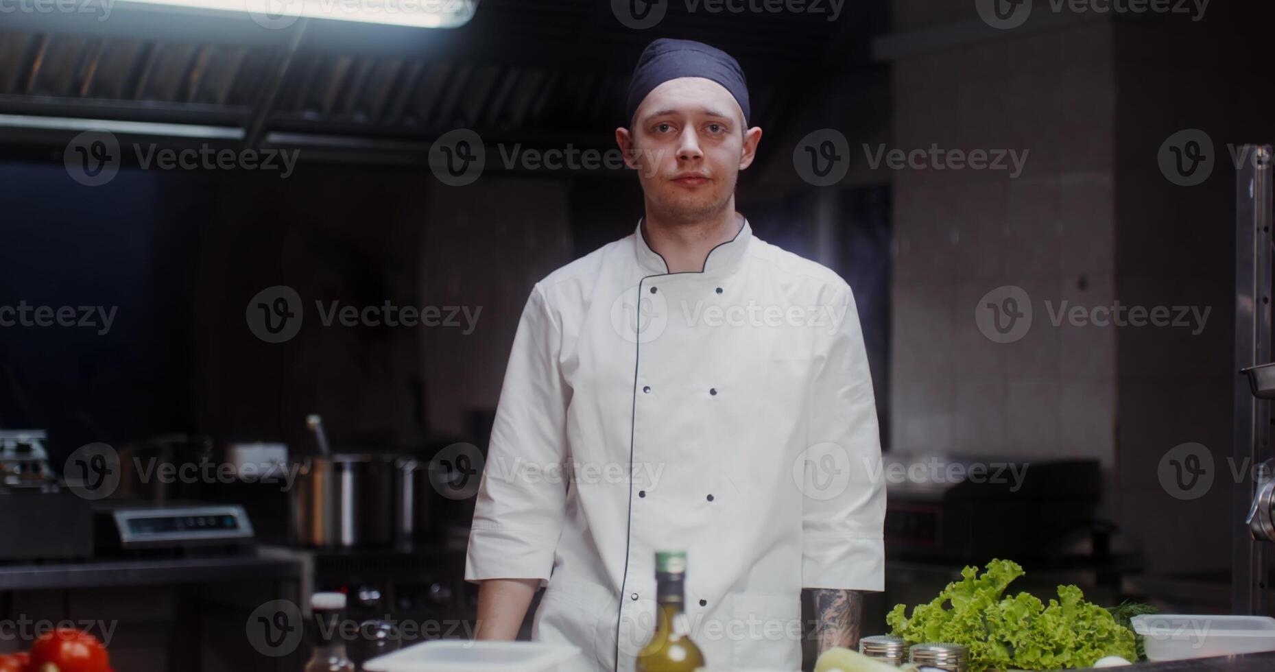 A man in a chef's uniform with a headband smiles while looking at the camera in a well-equipped restaurant kitchen photo