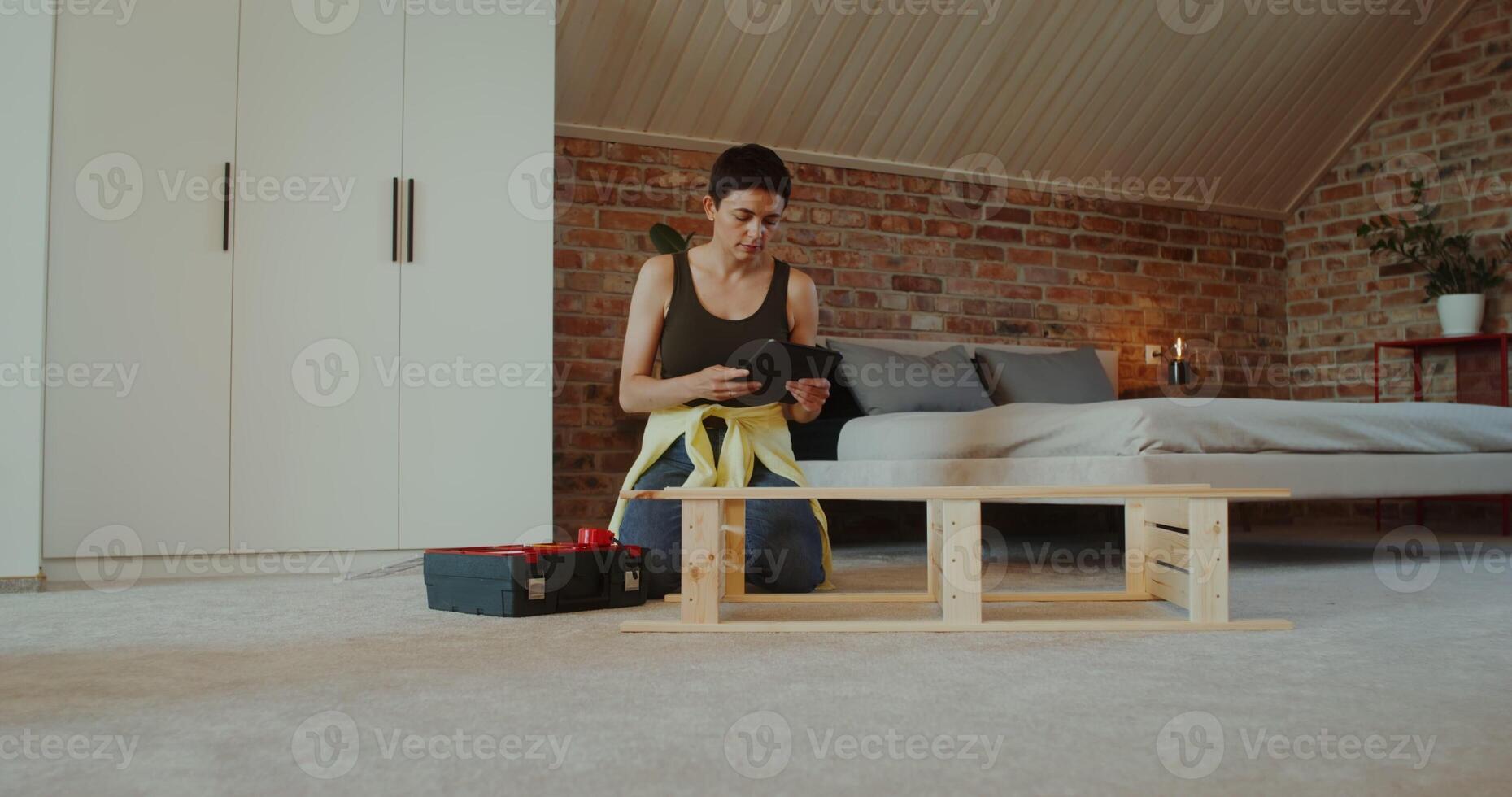 A young woman in casual clothes assembles a rack using a drill, sitting on the floor at home and looking at the tablet screen photo