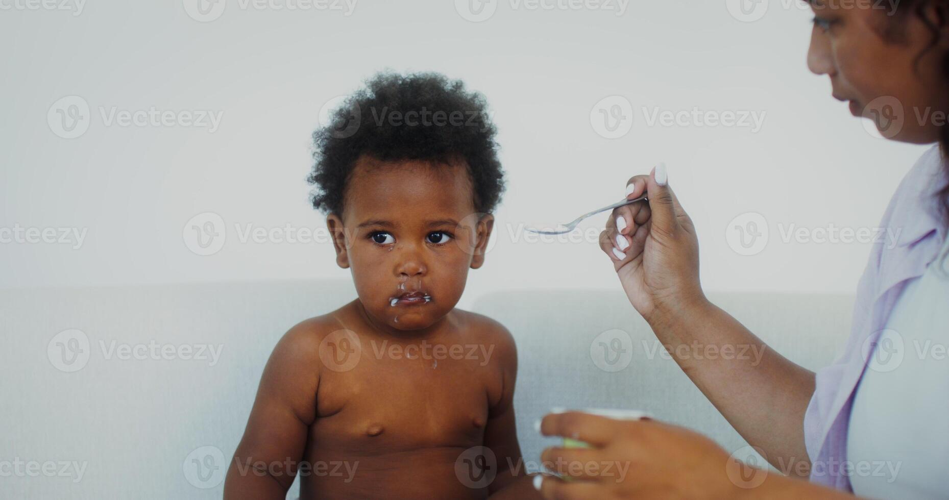 Mom feeds a small child of African-American appearance dressed in one diaper yogurt sitting on a sofa in a white interior photo