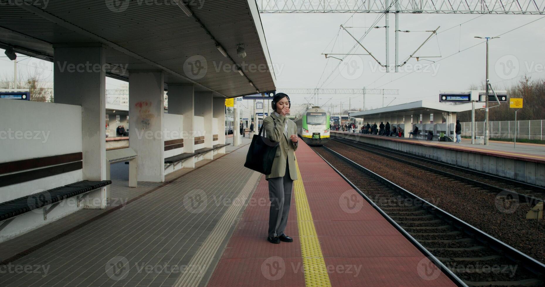 A woman in formal clothes and with headphones on her head drinks coffee from a disposable cup standing on the railway platform photo