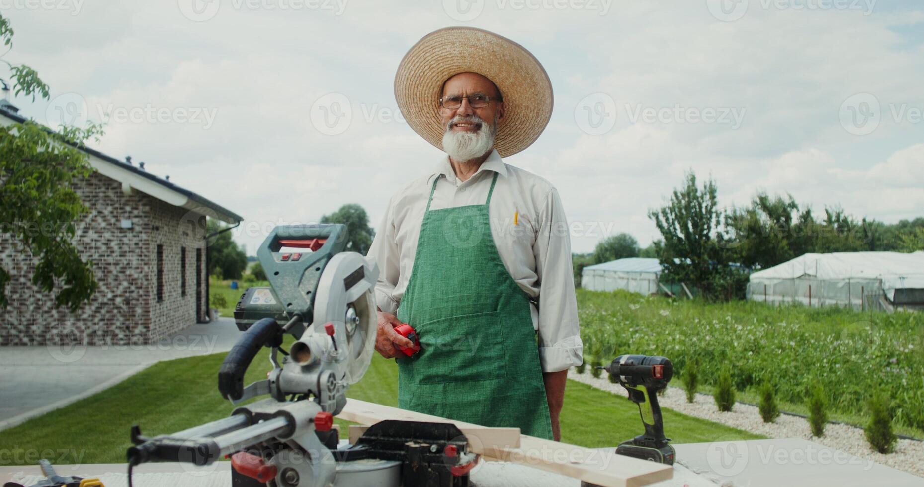 An elderly man with a gray beard, dressed in an apron and hat, smiles while looking at the camera standing in the backyard of his house near a miter saw photo