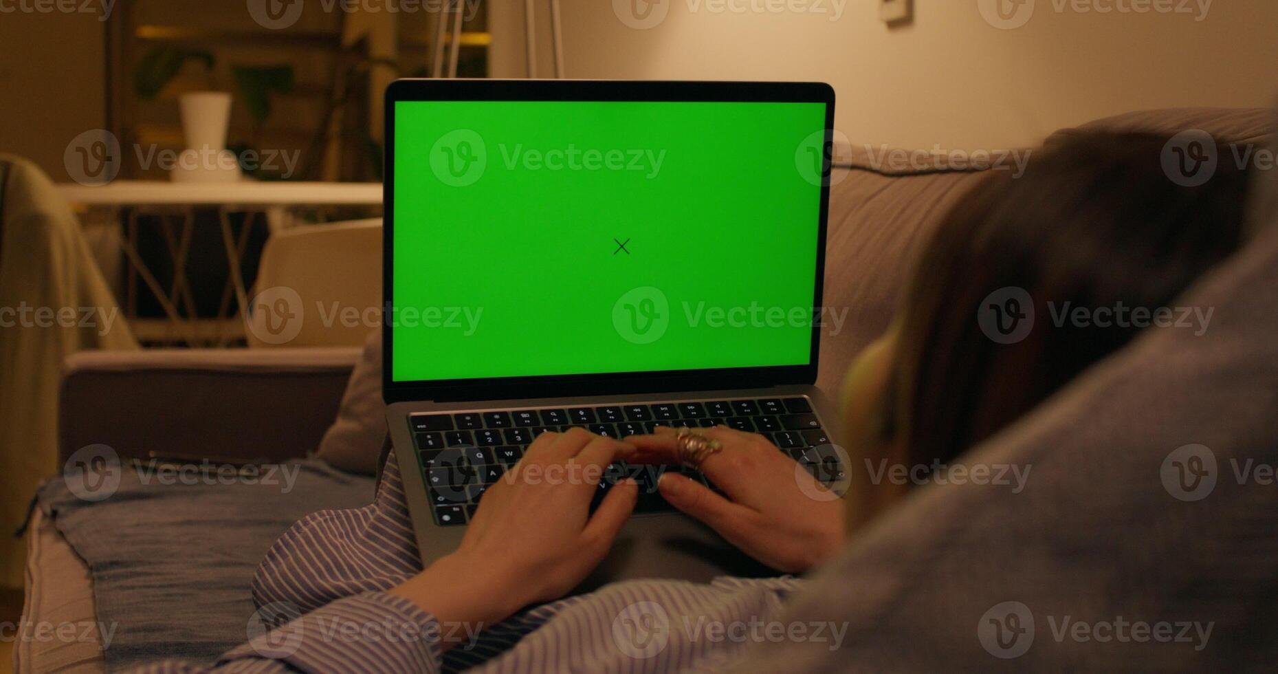 A woman typing on a laptop with a green screen lying on the couch in an apartment with dim lights, close-up of the screen, an unrecognizable person photo
