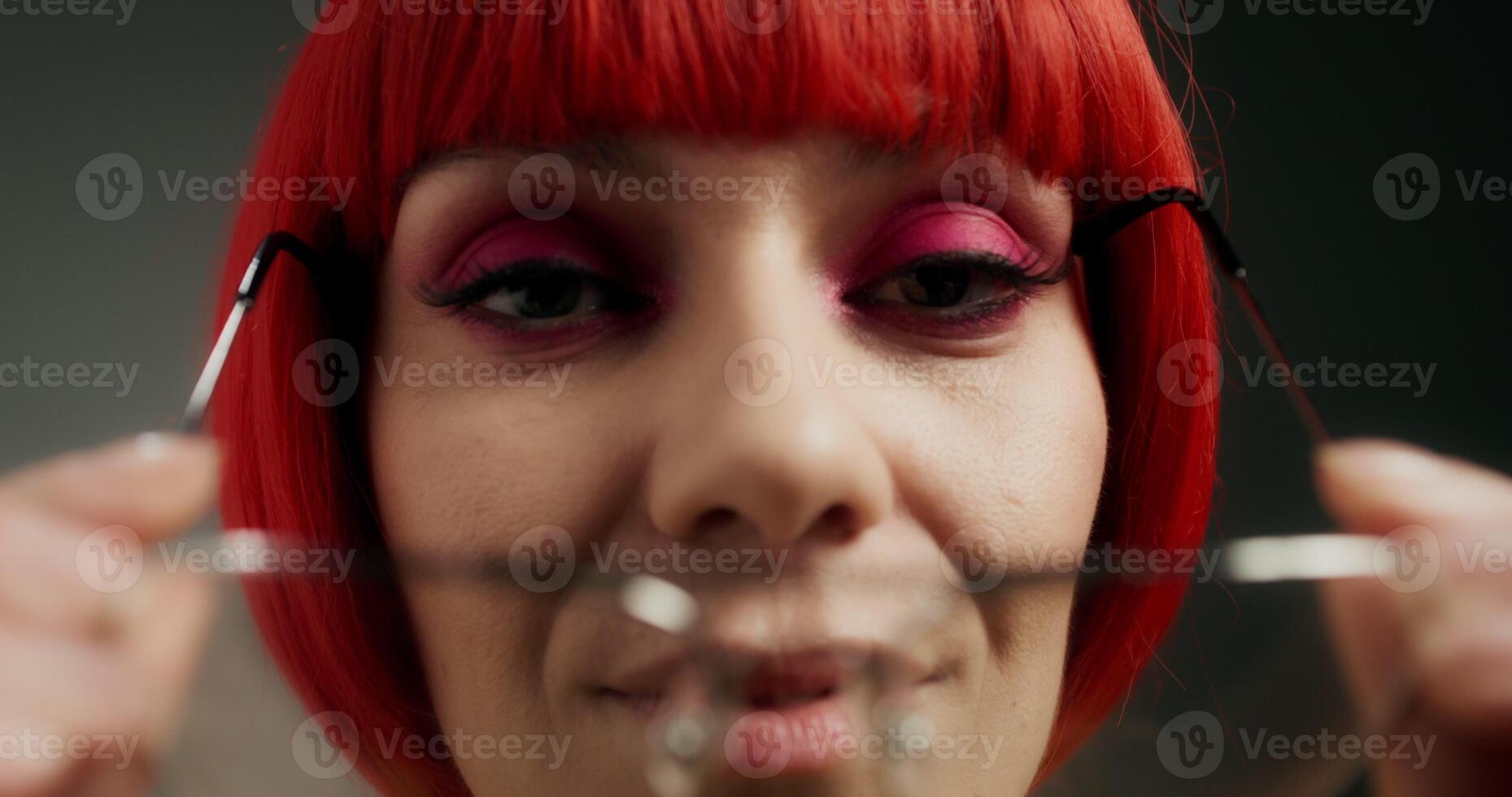 A woman with pink eyeliner and an orange wig puts on glasses and smiles looking at the camera, standing indoors, close-up of her face photo