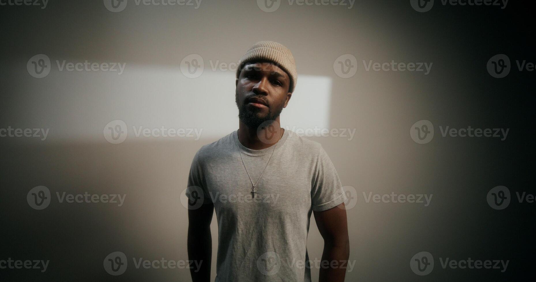 A stylish bearded man in a hat looks at the camera with a sad look standing on a white background in the studio photo