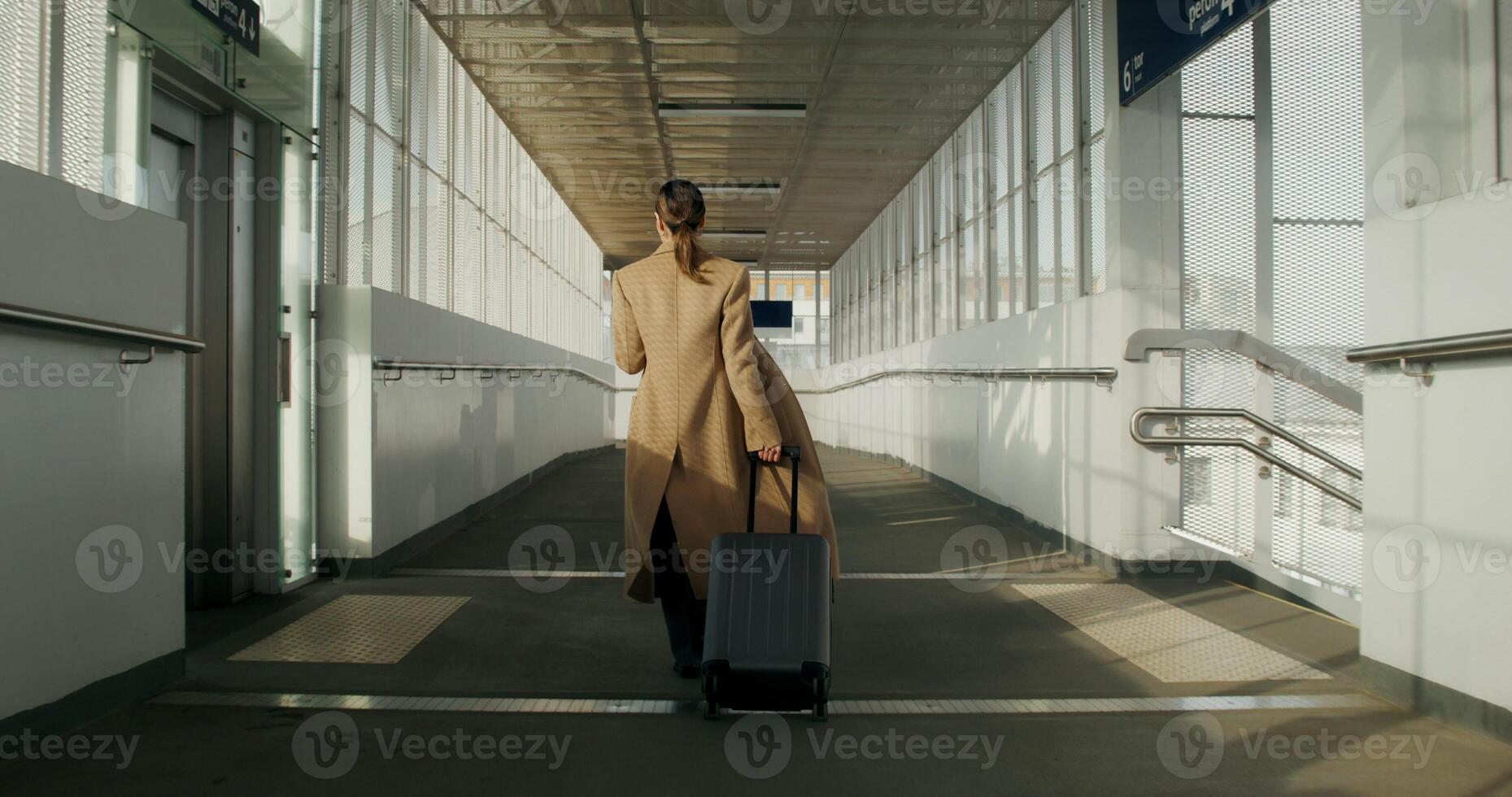 A woman in formal clothes and a coat uses a mobile phone walking along the corridor of the railway station, rear view photo