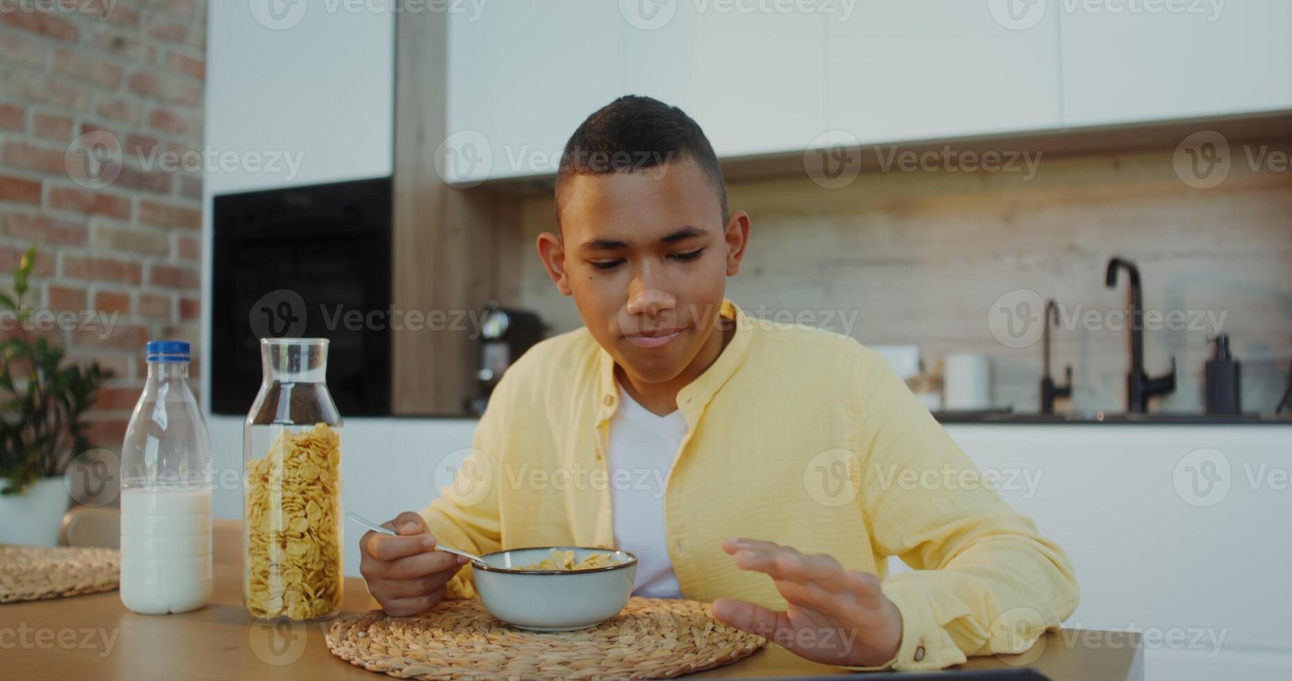 The boy has breakfast at home, he eats cereal with milk sitting at the table and looking at the tablet photo