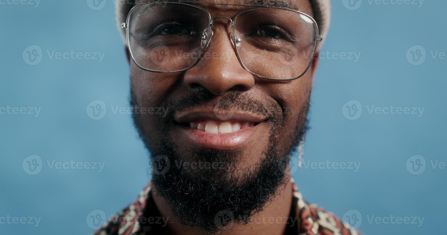 A bearded man with glasses and a hat, with an earring in his ear, smiling at the camera standing on a blue background in the studio, close-up of his face photo