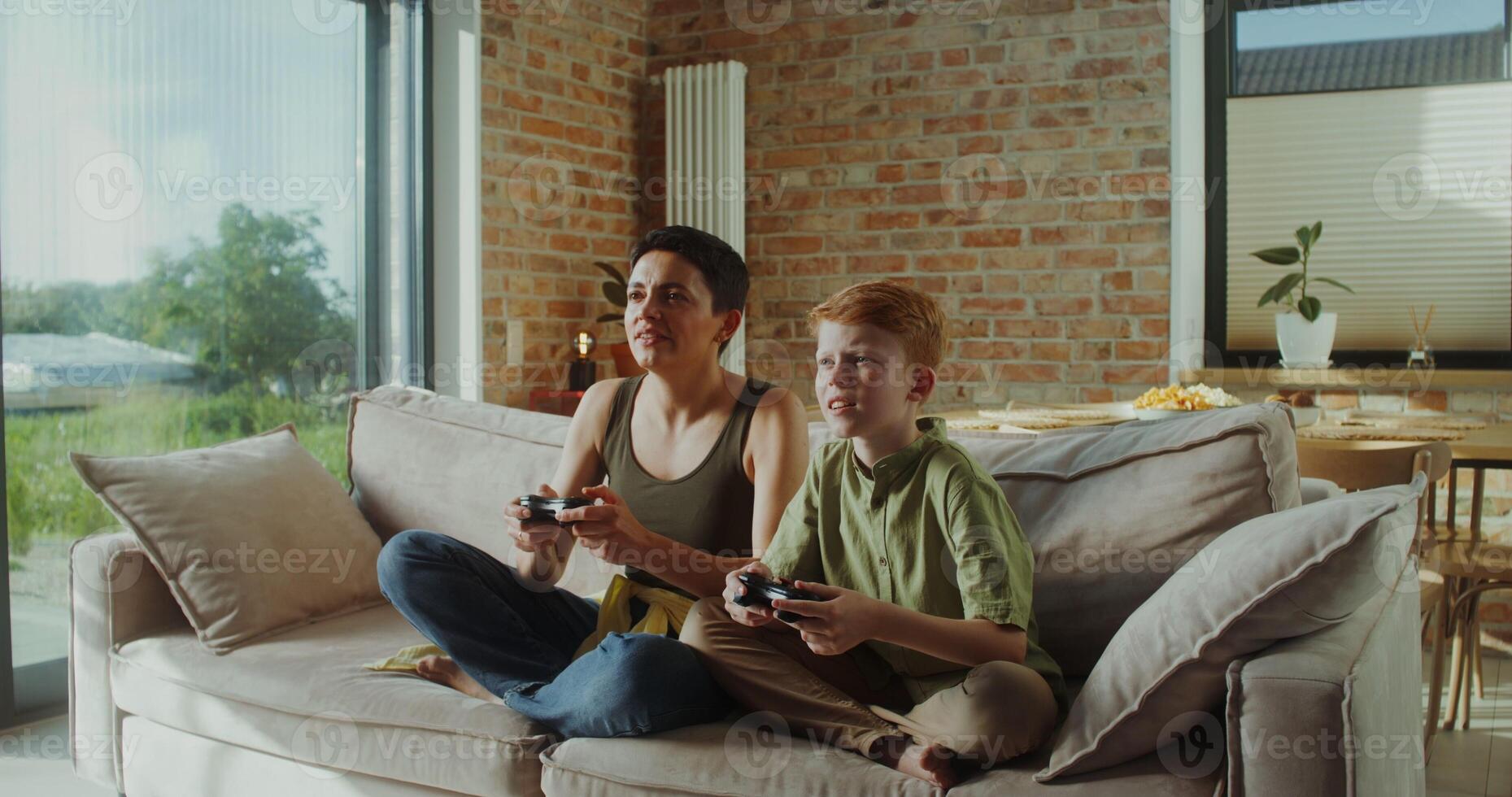 Mom and son of European appearance playing games using joysticks sitting on the couch in a stylish home interior. They rejoice in victory photo