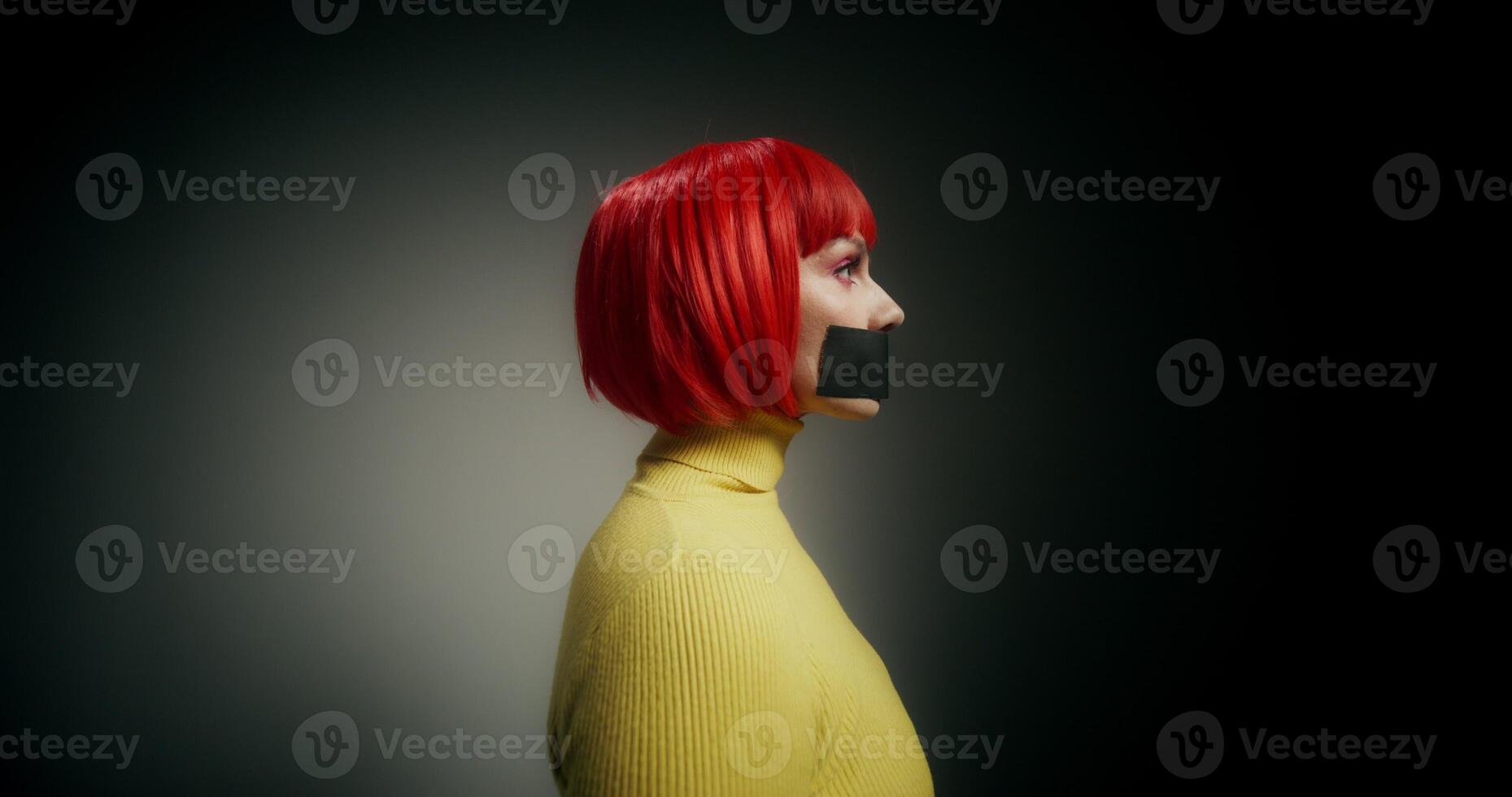 A woman in an orange wig with her mouth taped shut turns her head and looks at the camera with a frightened look photo