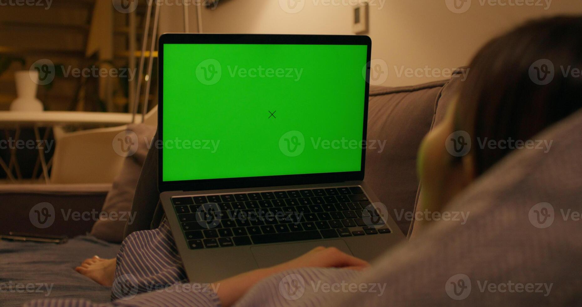 A woman uses a laptop with green screen lying on the couch in an apartment with dim lights, close-up of the screen, an unrecognizable person photo