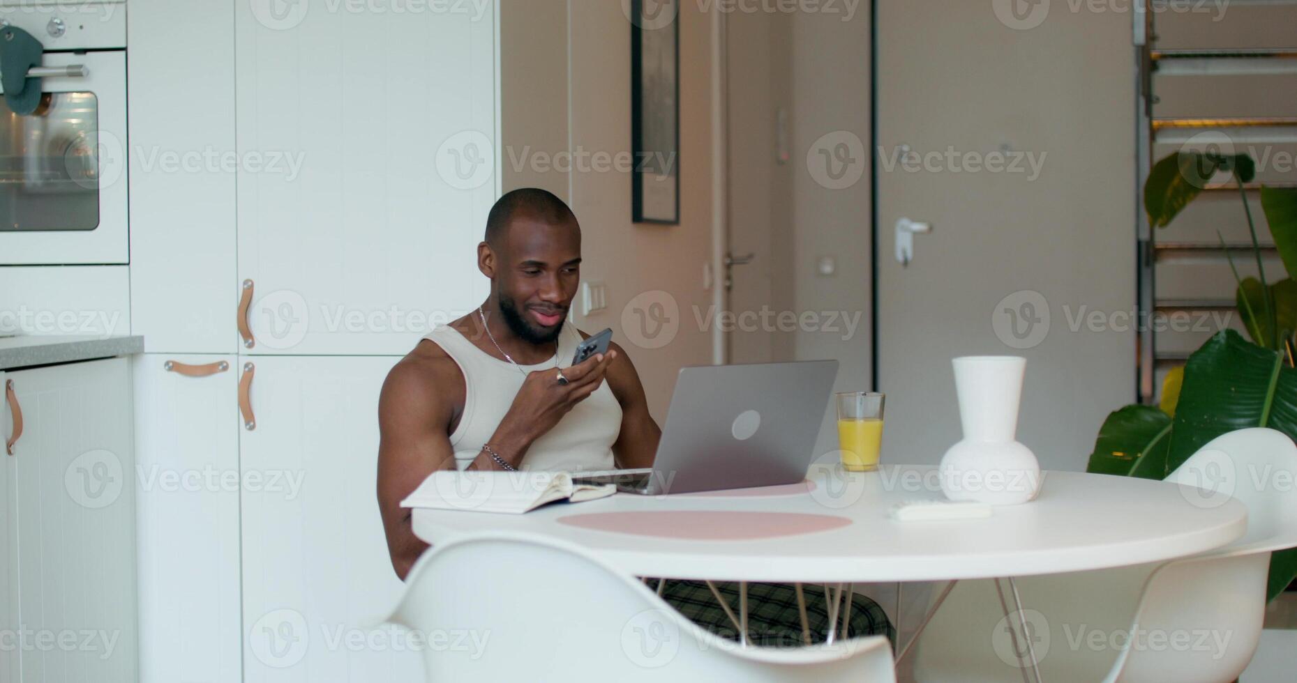 A man is smiling while recording a voice message on a mobile phone sitting at a table in the kitchen. An open laptop and a notebook in front of him photo