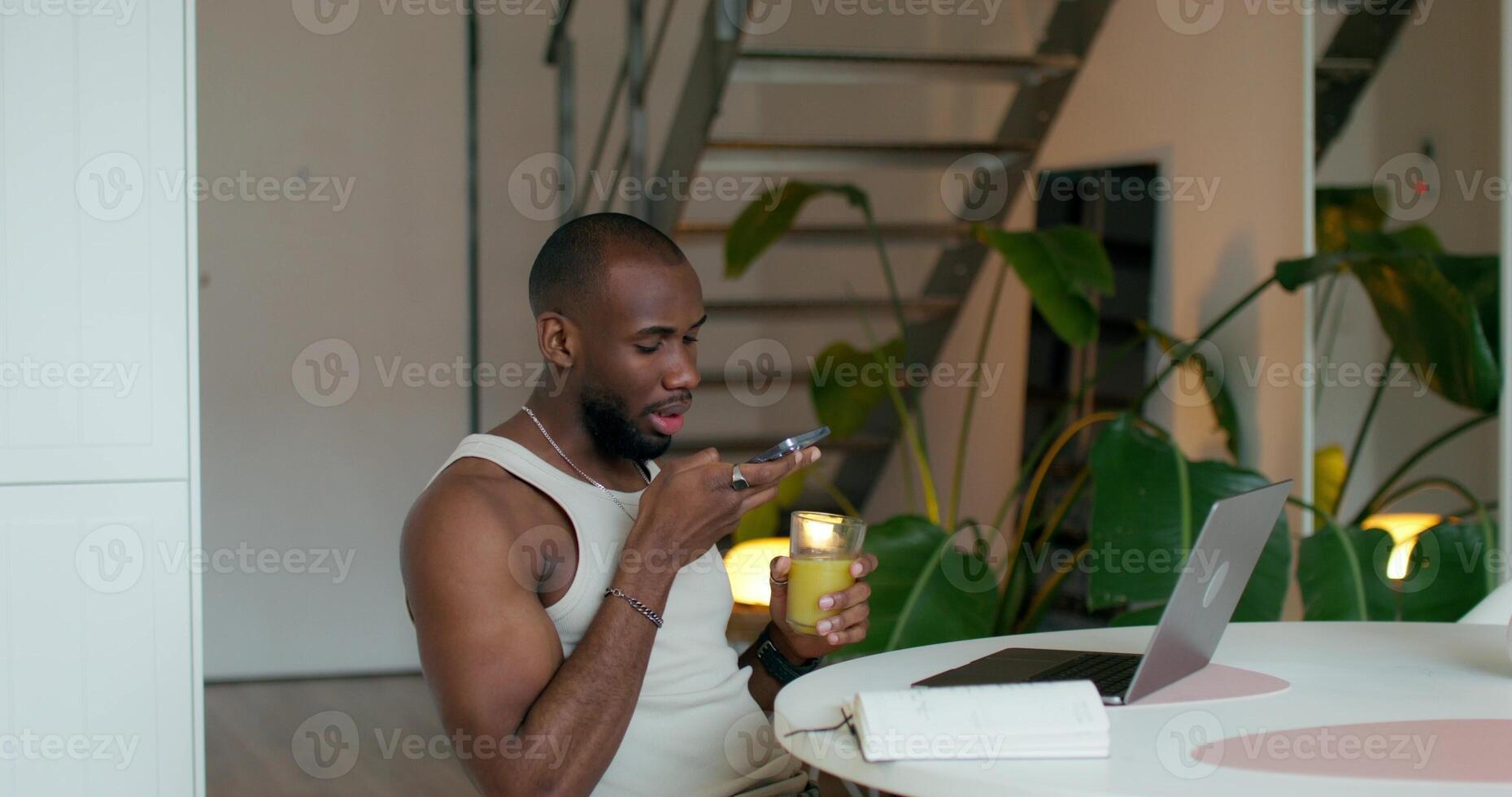 A man is recording a voice message on a mobile phone sitting at a table in the kitchen. An open laptop and a notebook in front of him photo