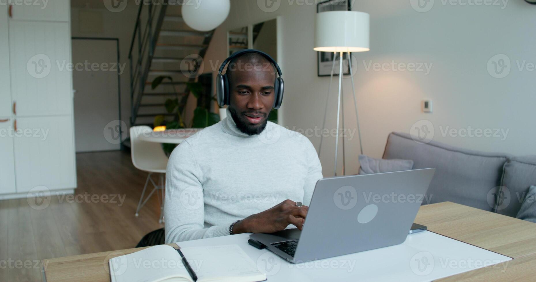 A man with headphones waves hello starting a chat on a laptop sitting at a table at home. He writes in a notebook. Remote work photo
