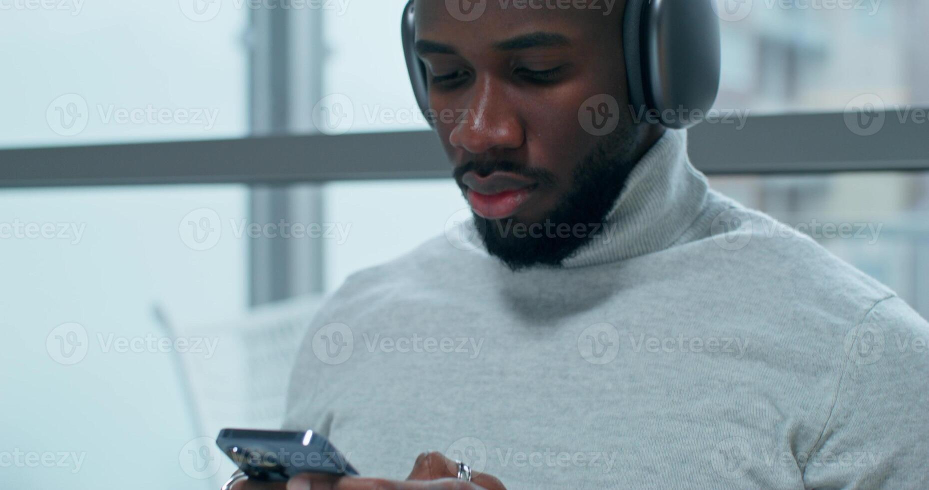 A man uses aA man wearing headphones uses a mobile phone sitting at a table indoors with a serious look. An open laptop is in front of him mobile phone indoors photo