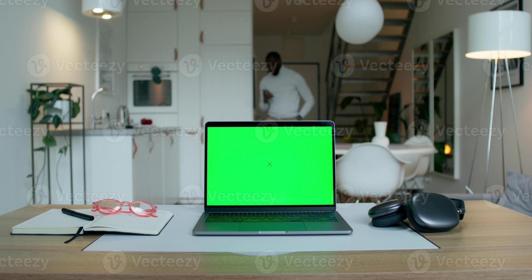 A laptop with a green screen is on the desktop at home. A man uses a mobile phone standing in the kitchen in the background photo