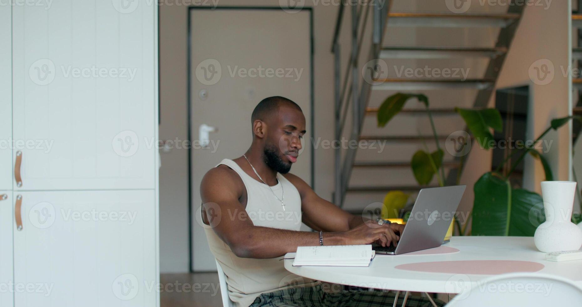 A man is smiling typing on a laptop and writing in a notebook while sitting at a table at home photo