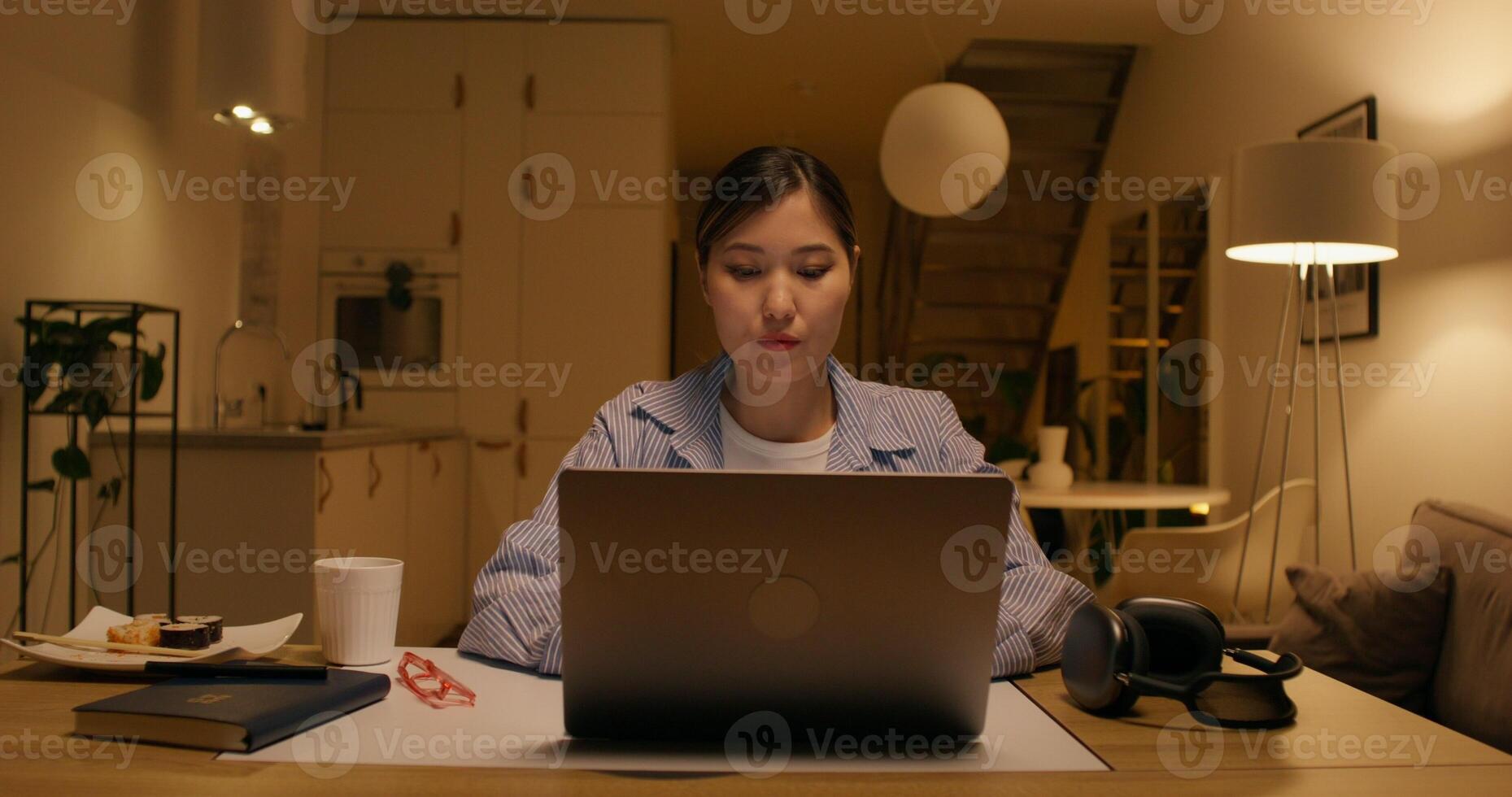 A woman is typing on a laptop and drinking coffee sitting at a table at home with a concentrated look, dimmed lights photo