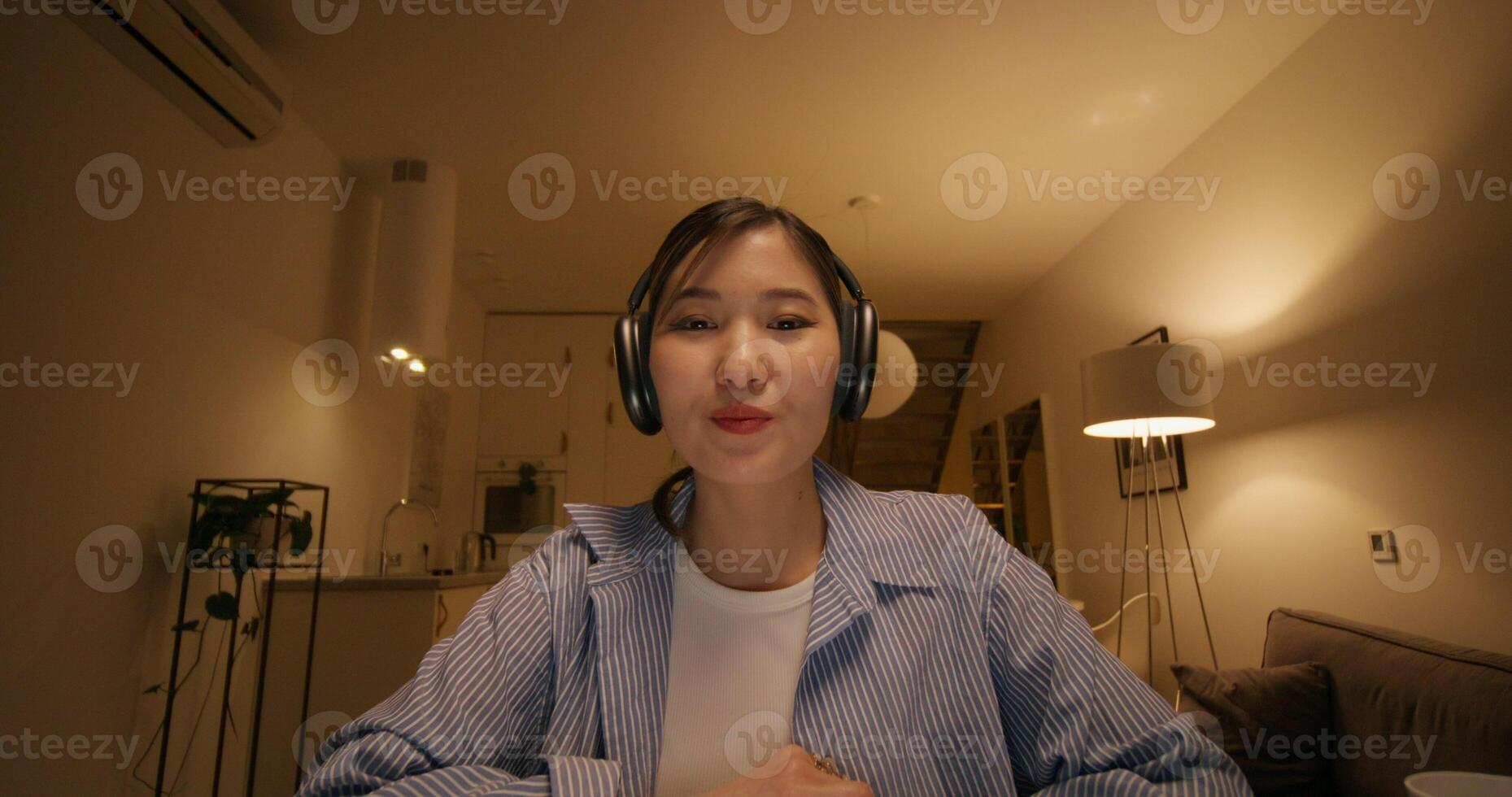 A woman with headphones is talking on a chat, looking at the camera as if at a laptop screen, sitting at a table in the dim light at home photo