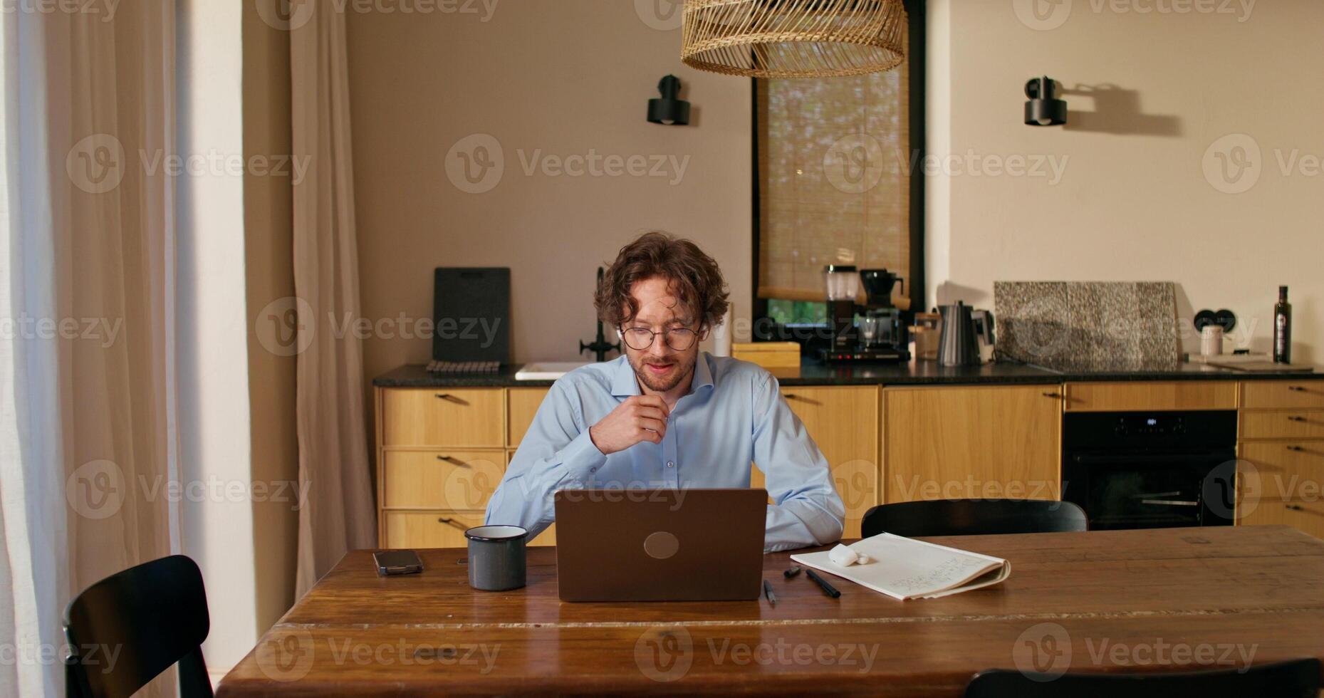 A man with glasses and earbuds is talking on a chat on a laptop and drinking coffee sitting at a table in the kitchen at home photo
