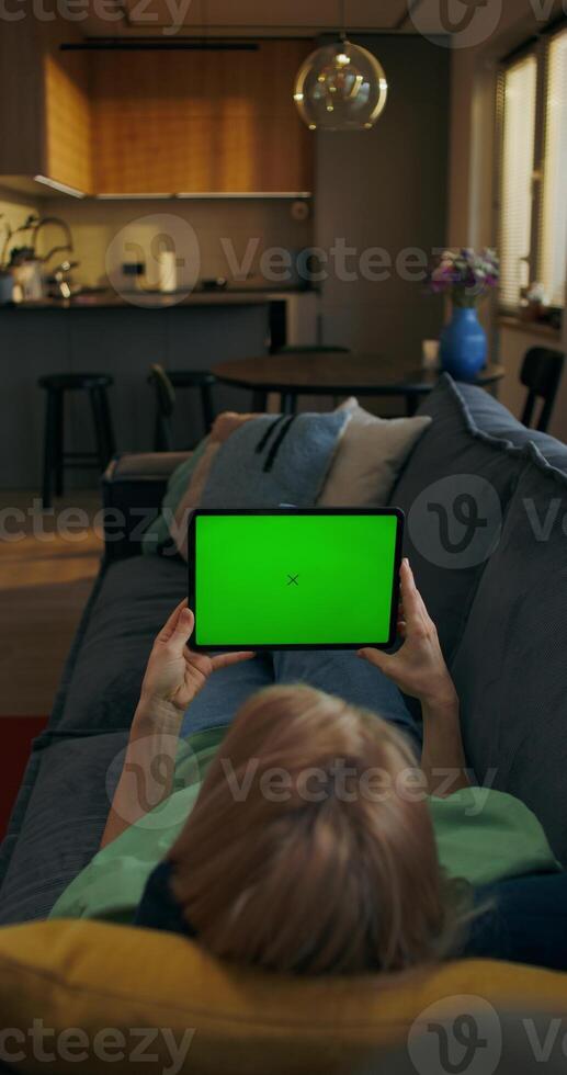 A woman is holding a tablet with green screen lying on the couch at home, an unrecognizable person photo